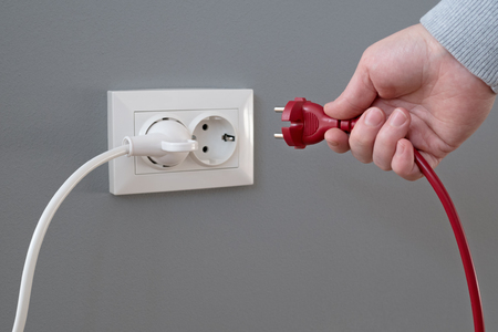 Hand plugging in an electric cord into a white plastic socket or  european wall outlet on grey plaster wall. Closeup of a woman's hand inserting an electrical plug into a wall socket. Daylight.の写真素材
