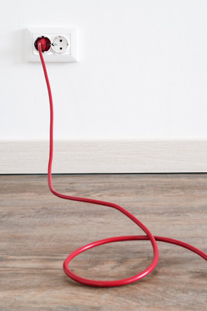 Red power cord cable plugged into european wall outlet on white plaster wall with copy space....