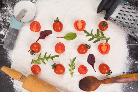 Whole and halved tomatoes on flour, arugula and basil. Tools for pizza preparation, rolling, spoon, cutter pizza, graterの写真素材