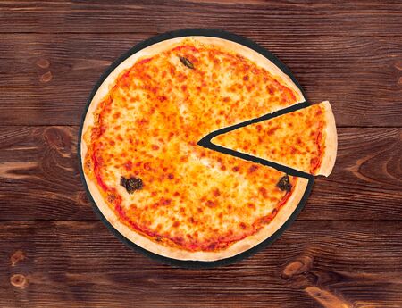 Margherita pizza with mozzarella, bocconcini and basil leaves, with a slice slightly removed on slate black platter which is on wooden background, top viewの写真素材