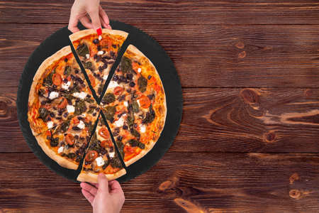 Young people taking slices of tasty pizza with cherry tomatoes, spinach, mozzarella, feta, kalamata olive and mushrooms from the slate black platter which is on wooden rustic table, space for textの写真素材