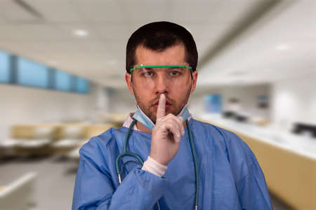 Doctor wearing medical coat, stethoscope, mask, gloves and goggles, protective against Coronavirus or Covid-19 in the hospital asking to be quiet with finger on lipsの写真素材
