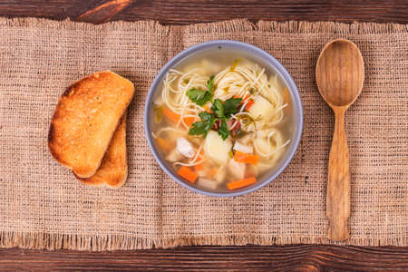 Vegetable soup with chicken pieces, vegetables and fresh herbs in bowl, wooden spoon and slices of toast on a textured burlap. Top viewの写真素材