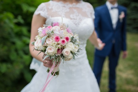 Wedding couple holding hands on summer green background with Rich bunch of pink peonies and lilac eustoma roses flowers. Wedding concept with bouquetの写真素材