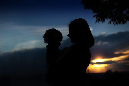 Silhouette of mother with her baby against the sunset outdoor. Mom and her toddler son are hugging on the sundown sky backgroundの写真素材