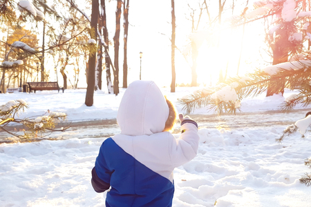 Year-old laughing baby boy in warm snowsuit walking in the winter park with a white snow. First winter and first toddler steps on the snow. Silhouette little child walking on a winter sunny dayの写真素材