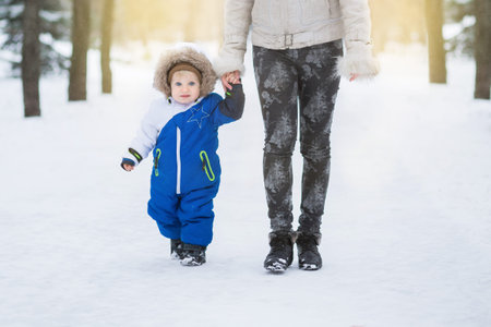 Mother and little toddler boy walking in the winter forest and having fun with snow. Family enjoying...