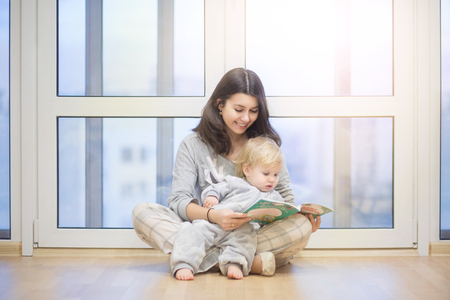 Happy young mother reading a book her year-old baby boy sitting near a window. Sunrise and sunshine, happy family lifestyle conceptの写真素材