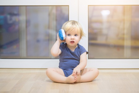 Portrait of a beautiful baby boy on the floor with music phone toys. 1,5 year-old child playing with educational cup toys at home. Little blond kid with blue eyes is playing with toy at home indoorsの写真素材