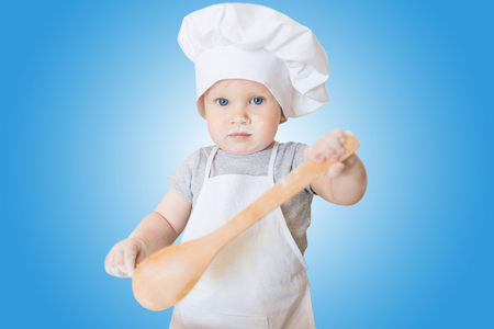 Little baby scullion is kneading dough in an apron and chef's hat isolated on blue background. Cooking child lifestyle conceptの写真素材