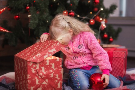 Adorable little girl opening a magical Christmas gift near a Christmas tree in cozy living room in winter. New year kid conceptの写真素材