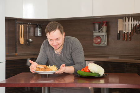 Man chooses between fast food and vegetables in the kitchen. Harmful - burger and fries unhealthy and wholesome food, lifestyle concept.の写真素材