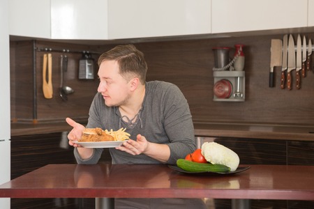 Man chooses between fast food and vegetables in the kitchen. Harmful - burger and fries unhealthy and wholesome food, lifestyle concept.の写真素材