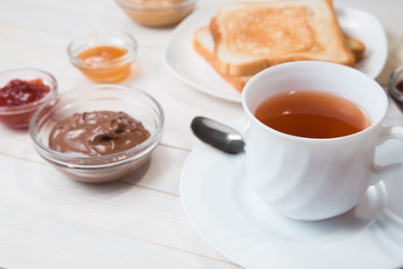 A white cup of black tea with Sandwiches or toasts with peanut butter, chocolate paste and strawberry, currant and apricot jelly or jam on white wooden table, close up, Breakfast conceptの写真素材