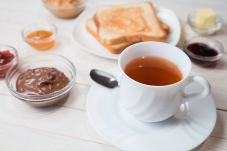 A white cup of black tea with Sandwiches or toasts with peanut butter, chocolate paste and strawberry, currant and apricot jelly or jam on white wooden table, close up, Breakfast conceptの写真素材