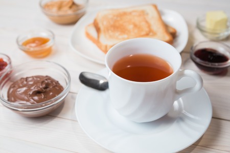 A white cup of black tea with Sandwiches or toasts with peanut butter, chocolate paste and strawberry, currant and apricot jelly or jam on white wooden table, close up, Breakfast conceptの写真素材