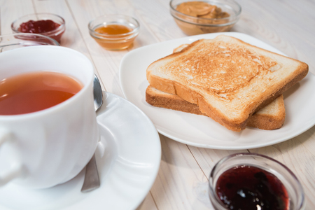 A white cup of black tea with Sandwiches or toasts with peanut butter, chocolate paste and strawberry, currant and apricot jelly or jam on white wooden table, close up, Breakfast conceptの写真素材