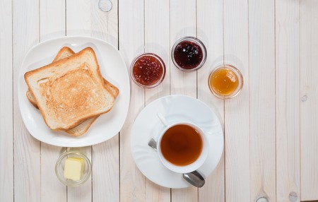 A white cup of black tea with Sandwiches or toasts with strawberry, currant and apricot jelly or jam on white wooden table, top view, flat lay, Breakfast concept with copy spaceの写真素材