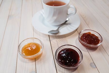 Breakfast with cup of tea and strawberry, currant and apricot jelly or jam on rustic wooden white background, close upの写真素材