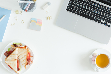 Overhead view of laptop, fresh sandwich, cup of green tea and mobile phone on white desktop table. Woman business and breakfast concept, top view and flat layの写真素材