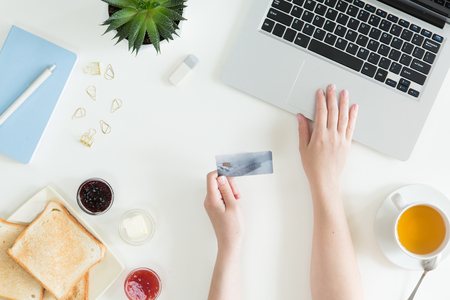 Top view of woman hands holding credit card, business lunch concept, online shopping, workspace with laptop, mobile phone, flowers and notebook, toast, flat lay.の写真素材
