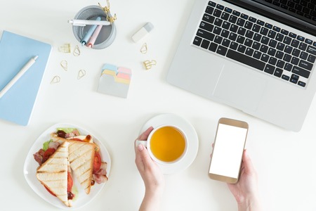 Top view of break time on white office table desk. Business lunch in office, top view of sandwich, cup of green tea on white desk with laptop, mobile phone and notepad, flat lay.の写真素材