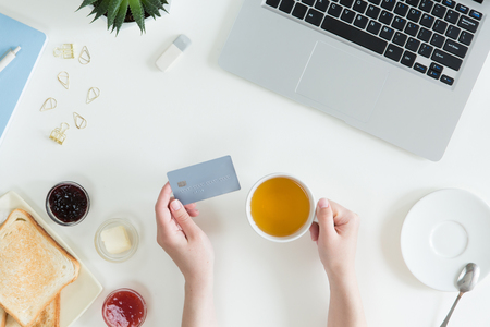 Top view of woman hands holding credit card, business lunch concept, online shopping, workspace with laptop, mobile phone, flowers and notebook, toast, flat lay.の写真素材