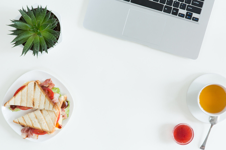 Overhead view of laptop, fresh sandwich, cup of green tea and mobile phone on white desktop table. Woman business and breakfast concept, top view and flat layの写真素材