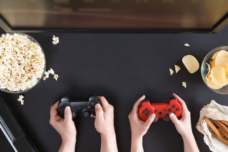 Man and woman playing video games together, top view of gamer accessorises and snacks frame, flat lay on black background. Joystick and gamepad, keyboard, game console, beer, chips and popcorn.の写真素材