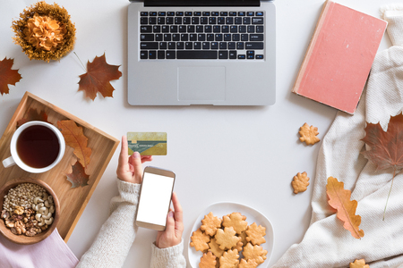 Top view of woman hands holding credit card on autumn baclground, business concept, online shopping, workspace with laptop, mobile phone, autumn leaves and flowers and notebook, cookies, flat lay.の写真素材