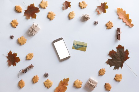 View from above of autumn background with mobile phone, credit card, autumn leaves, cookies, vintage book on white workspace. Top view and flat layの写真素材