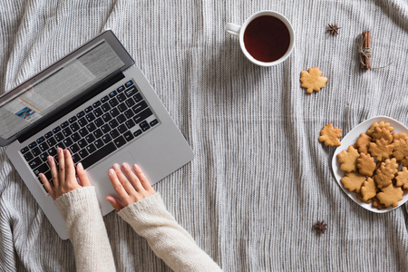 Overhead view of morning tea mug composition with autumn leaves, cookies on knitted background, flat lay. Cozy breakfast on woman workspace with laptop and vintage book on wrap.の写真素材
