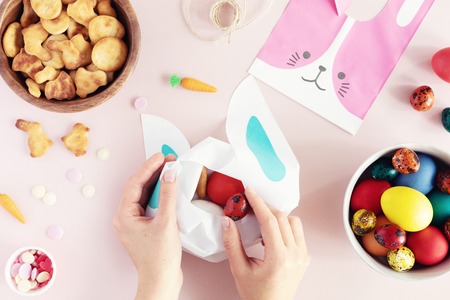 Top view and flat lay of Easter painted eggs, cookies, sprinkles and candies arranged on pink background. Woman hands fill a bag or basket with sweet giftsの写真素材