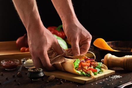 Shawarma sandwich in men hand on wooden rustic background. Gyro fresh roll of lavash or pita bread with grilled chicke, lettuce salad, cheese and vegetables. Tasty shawarma - middle eastern snack.の写真素材