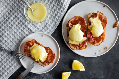 Eggs Benedict- toasted English bread, parm ham, poached eggs with hollandaise sauce and lemon, view from above on gray backgroundの写真素材