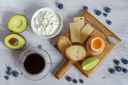 A healthy and nutritious breakfast: a bowl of cottage cheese, hard cheese, boiled egg, baguette slices with avocado and cup of hot coffee or tea.の写真素材