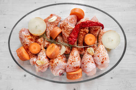 Raw chicken drumsticks with herbs, carrots, chopped onions in glass roasting tray on light wooden backgroundの写真素材