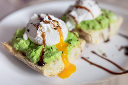 Poached egg on a slice of toast bread, with guacamole and dripping yolk, on a white plate