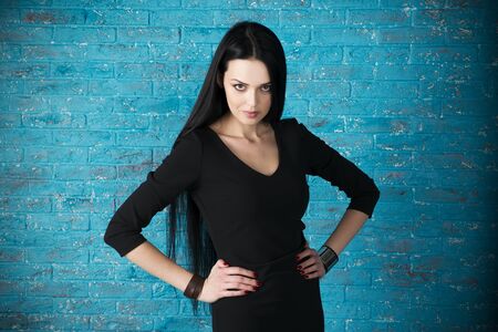 Beautiful young woman in a black dress posing against the backdrop of a blue brick wall. Half-lengthの写真素材