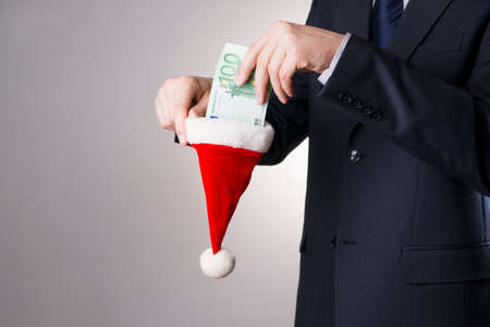 Businessman putting money in a Santa hat on gray backgroundの写真素材