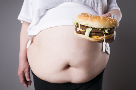 Fat women suffer from obesity with big hamburger in hand, junk food concept on a gray backgroundの写真素材