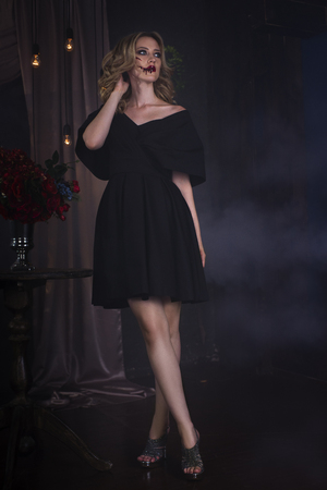 Beautiful young blonde woman in black dress with halloween make up and bloody face art, vintage interiorの写真素材