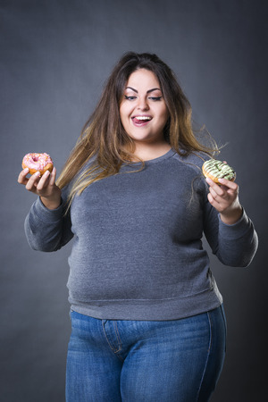 Happy beautiful young caucasian plus size model posing with donuts on a gray studio background, fast food and unhealthy nutrition conceptの写真素材