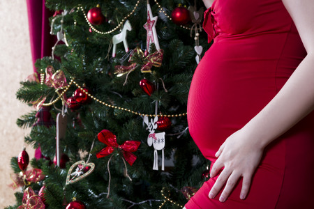 Young pregnant woman in red dress posing on the background of Christmas tree, holiday concertの写真素材
