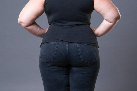Fat female buttocks in blue jeans, overweight woman body closeup, gray studio background