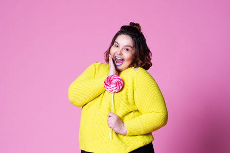 Cheerful plus size model with big lollipop, fat woman in yellow jumper on pink background, body...