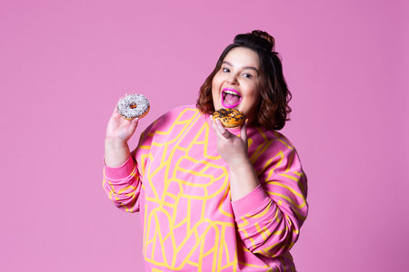 Cheerful plus size model eating doughnuts, fat woman overeats on pink background, unhealthy...