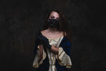 Beautiful woman in renaissance dress, face mask and gloves on abstract dark background, old and new conceptの写真素材