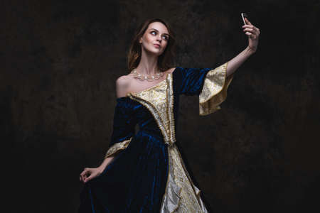 Beautiful woman in renaissance dress taking selfie on smartphone, old and new concept, abstract dark backgroundの写真素材