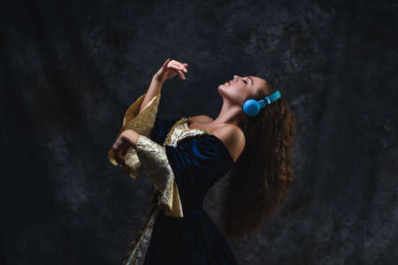 Beautiful woman in renaissance dress listens to music in wireless headphones on abstract dark background, old and new conceptの写真素材